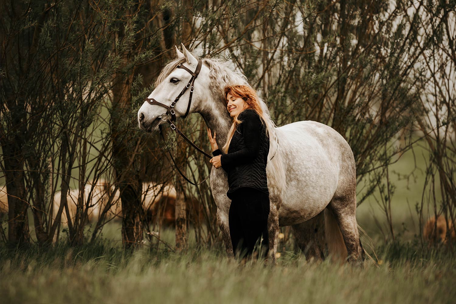 Eine Frau mit rotem Haar steht lächelnd neben einem weissen Pferd, das sanft in die Kamera blickt, eingebettet in eine natürliche Umgebung mit grünen Büschen.