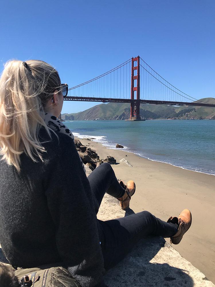 Eine Person sitzt am Ufer mit Blick auf die Golden Gate Bridge in San Francisco, von hinten gesehen, während sie den Ausblick geniesst.