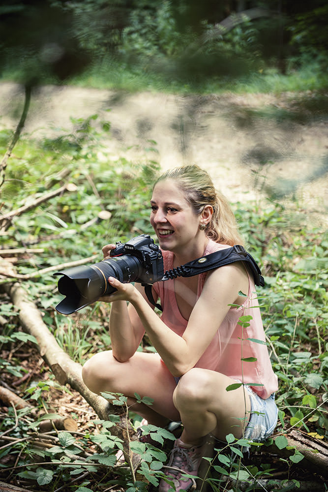 Lächelnde Fotografin hockt im Wald und hält eine professionelle Kamera, bereit zum Fotografieren.
