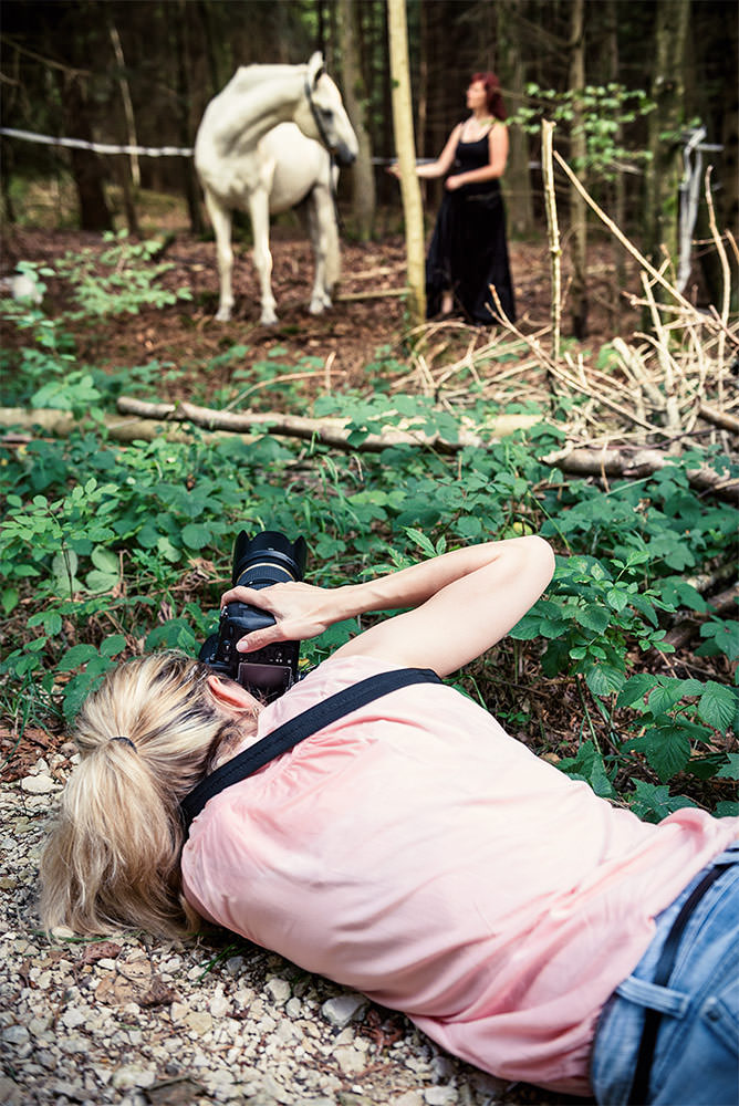 Pferdefotograf liegt auf dem Waldboden, um eine Frau in einem schwarzen Kleid neben einem weissen Pferd zu fotografieren.
