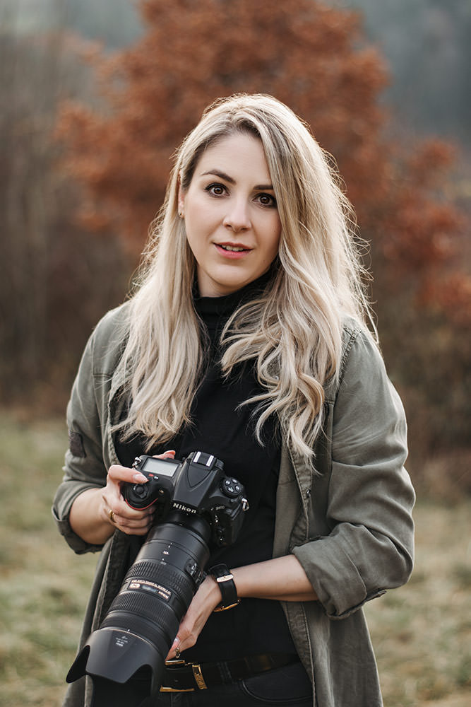 Pferdefotograf Schweiz mit langen blonden Haaren steht im Freien und hält eine Kamera, bekleidet mit einem schwarzen Rollkragenpullover und einer grünen Jacke vor einem Hintergrund aus herbstlichen Bäumen.