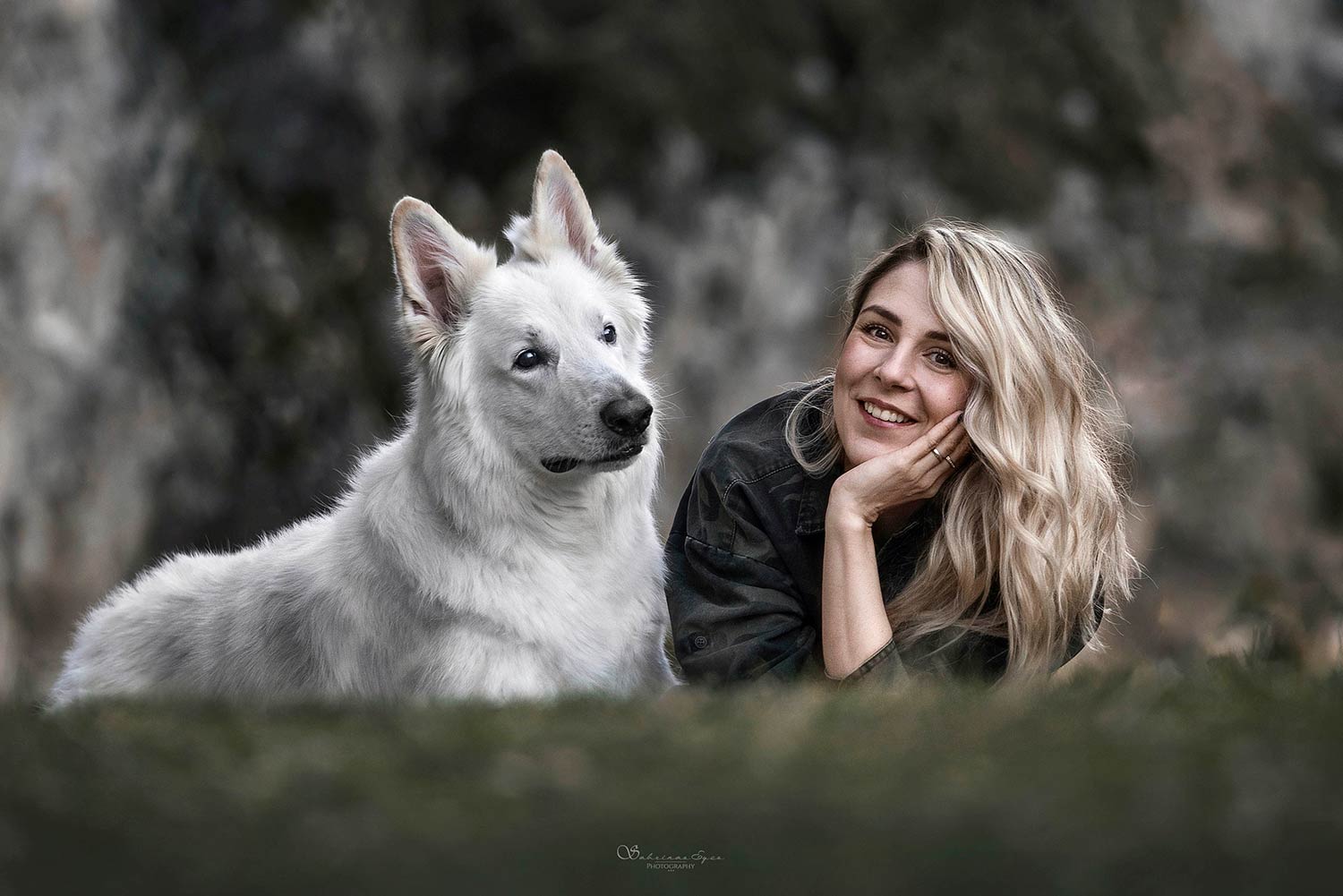 Eine lächelnde Frau liegt im Gras neben einem weissen Hund, beide blicken in die Kamera.