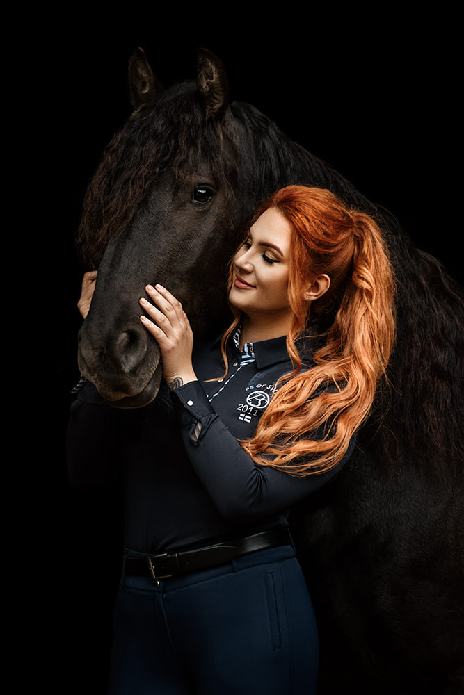 Eine Frau mit roten Haaren kuschelt liebevoll mit einem schwarzen Pferd bei einem Fotoshooting vor einem schwarzen Hintergrund.
