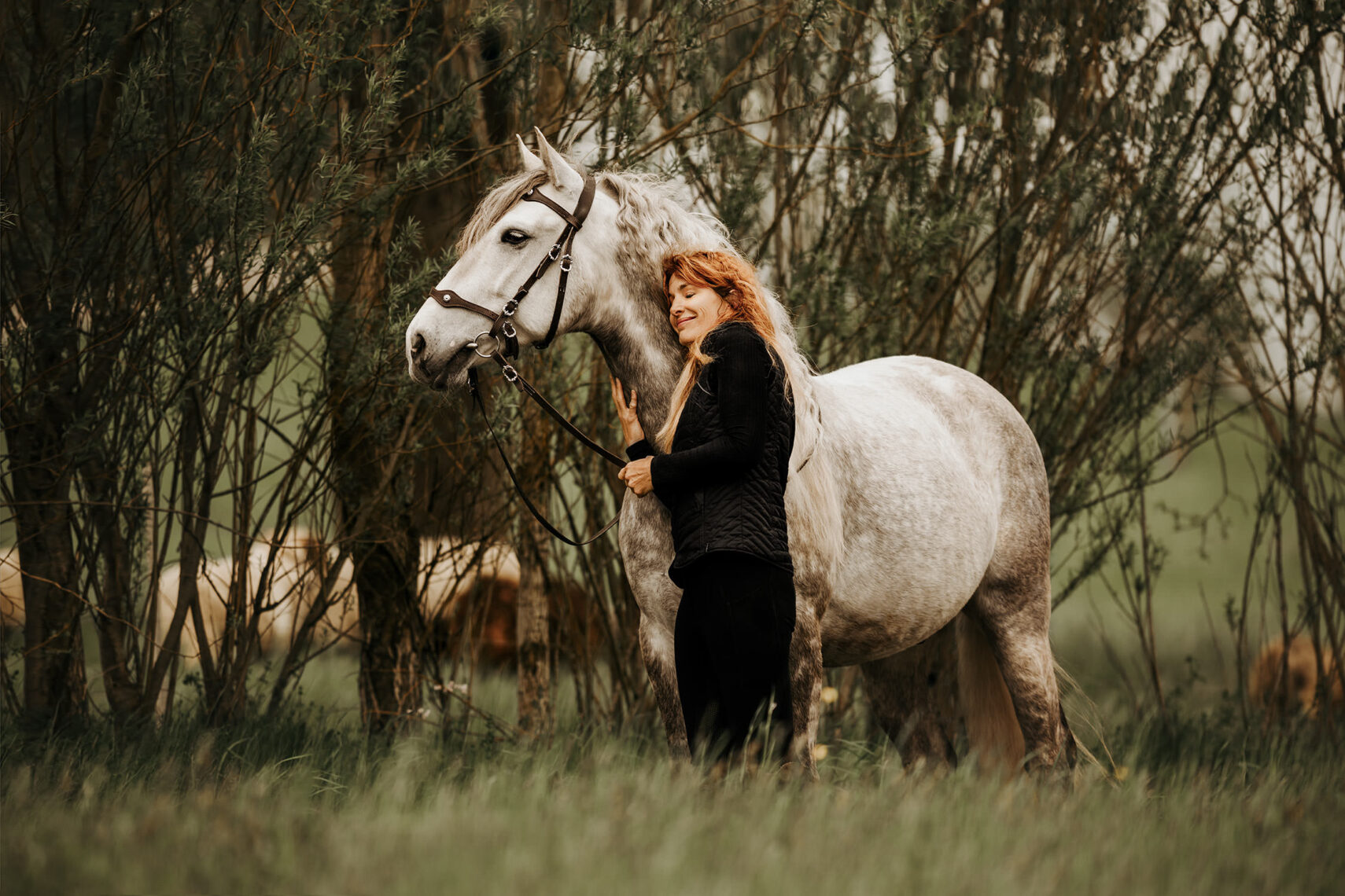Eine lächelnde Frau streichelt liebevoll ein weisses Pferd in einem grünen Buschwerk.