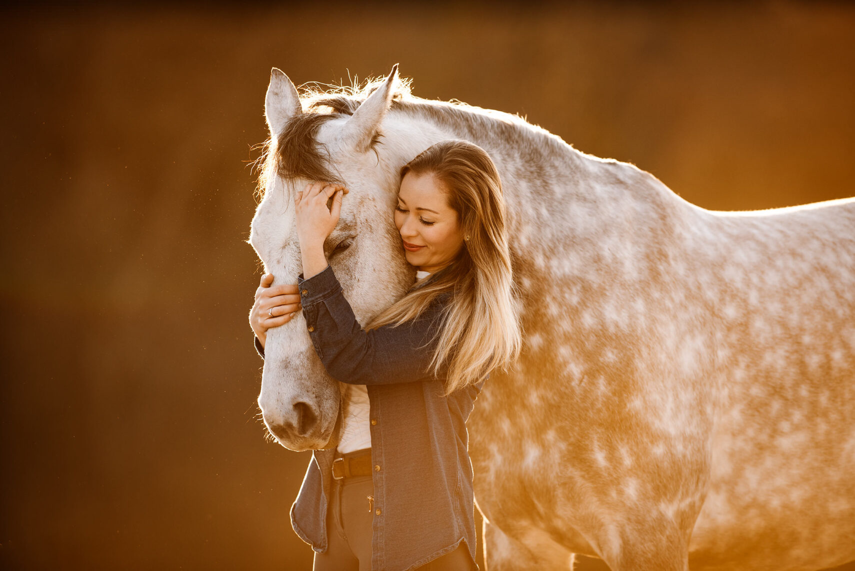 Eine innige Umarmung zwischen einer jungen Frau und einem apfelschimmel Pferd im goldenen Sonnenlicht.