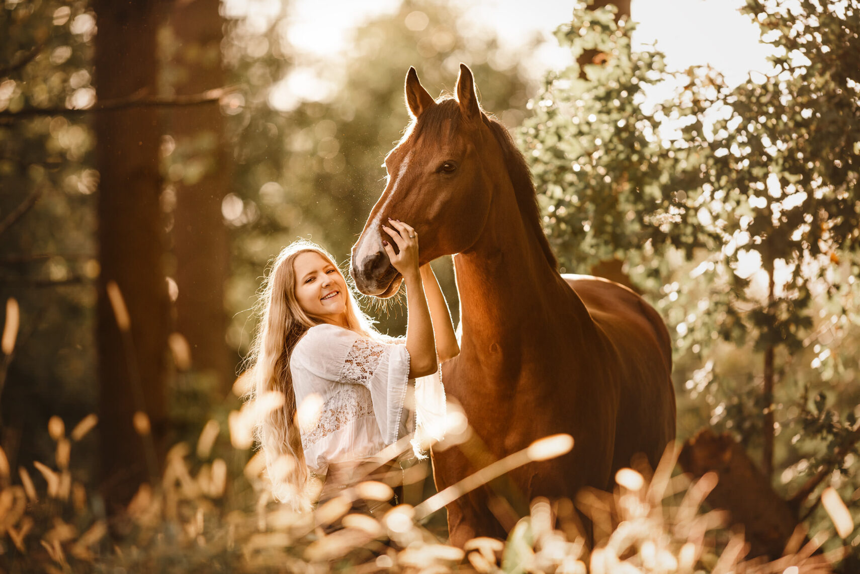 Eine junge Frau lächelt, während sie sanft das Gesicht eines Pferdes berührt, umgeben von der warmen Beleuchtung eines Waldes.