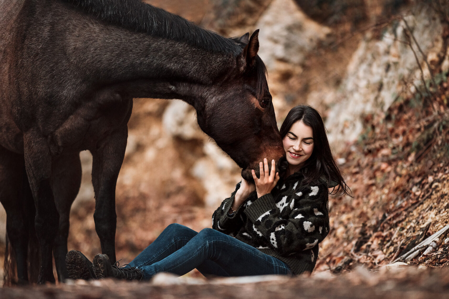 Eine junge Frau sitzt lächelnd im Herbstlaub, während sie zärtlich von einem Pferd berührt wird.