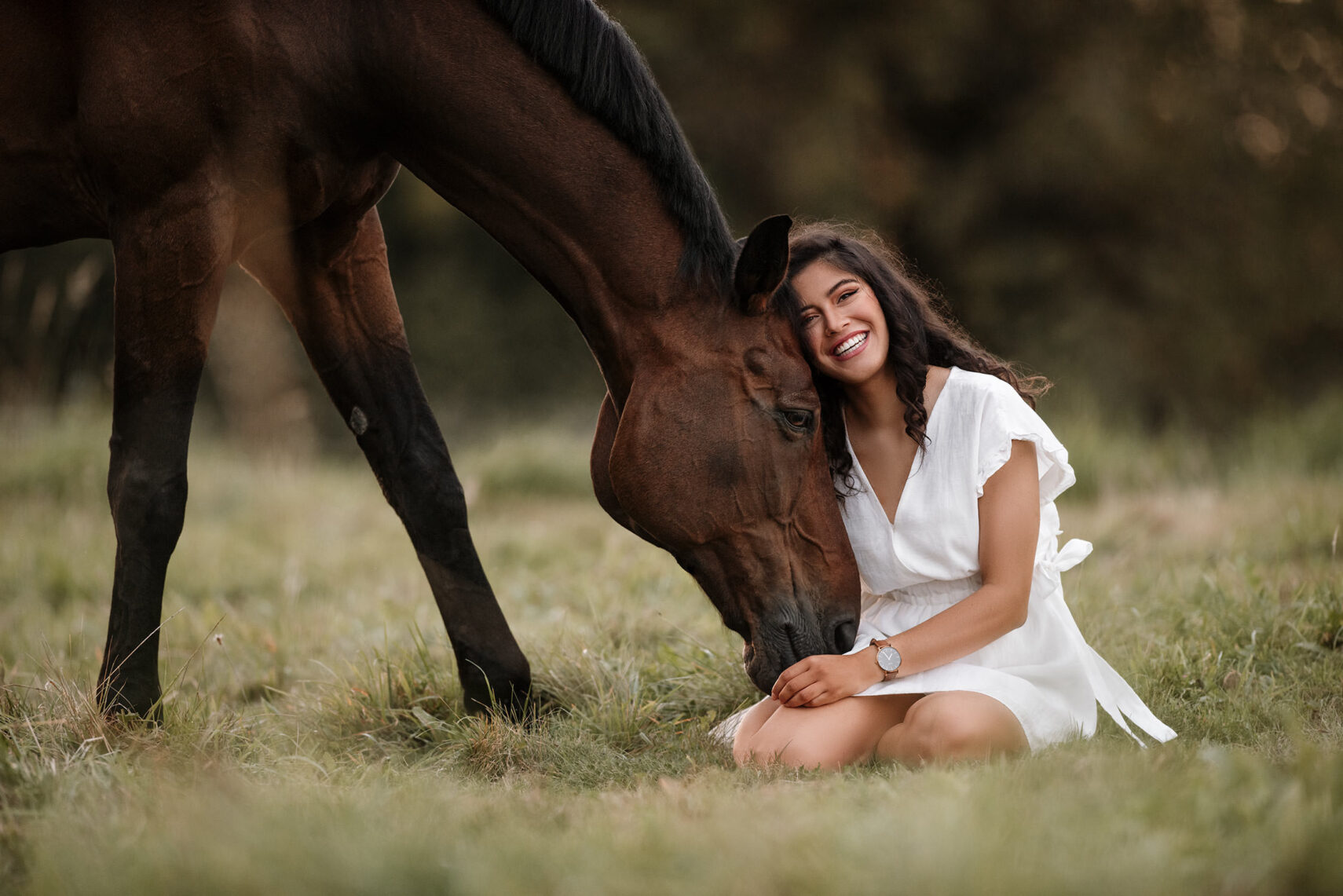 Ein strahlendes Lächeln teilt die junge Frau im weissen Kleid, während sie liebevoll von einem Pferd angestupst wird.
