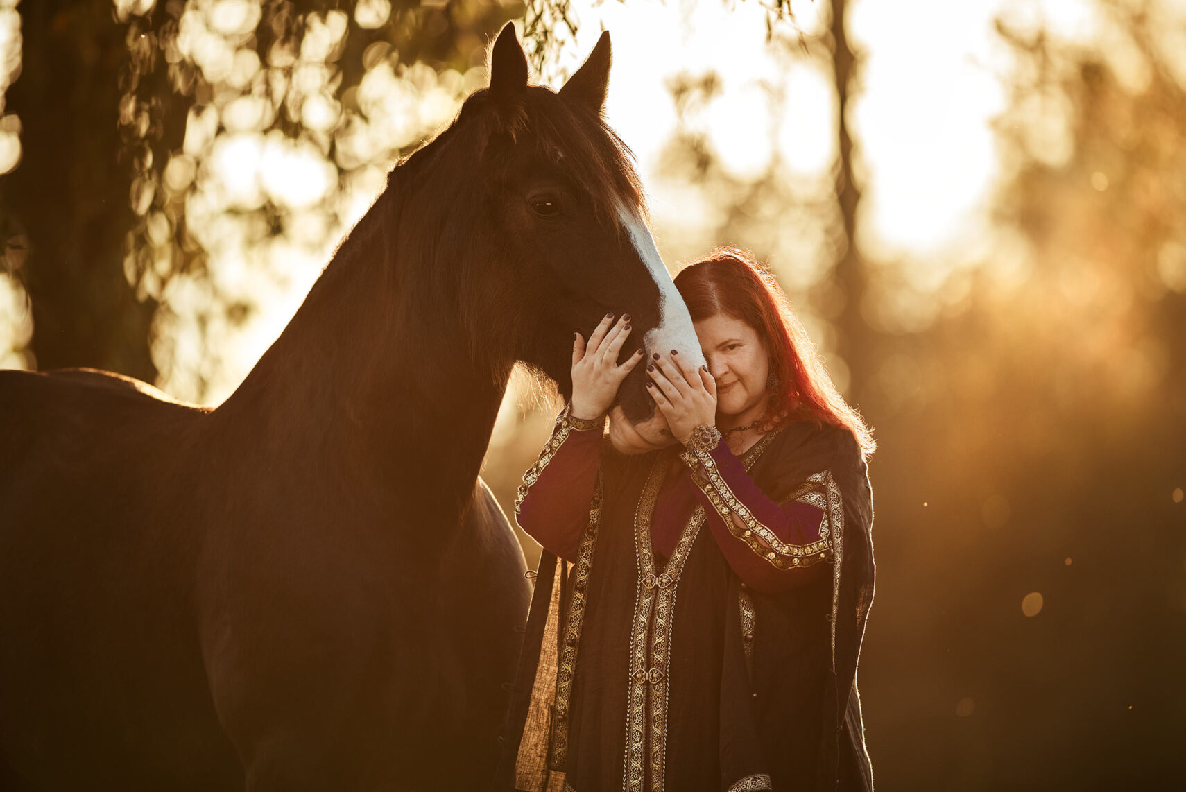 Eine Person mit rotem Haar umarmt liebevoll das Gesicht eines Pferdes im warmen Licht eines Sonnenuntergangs.