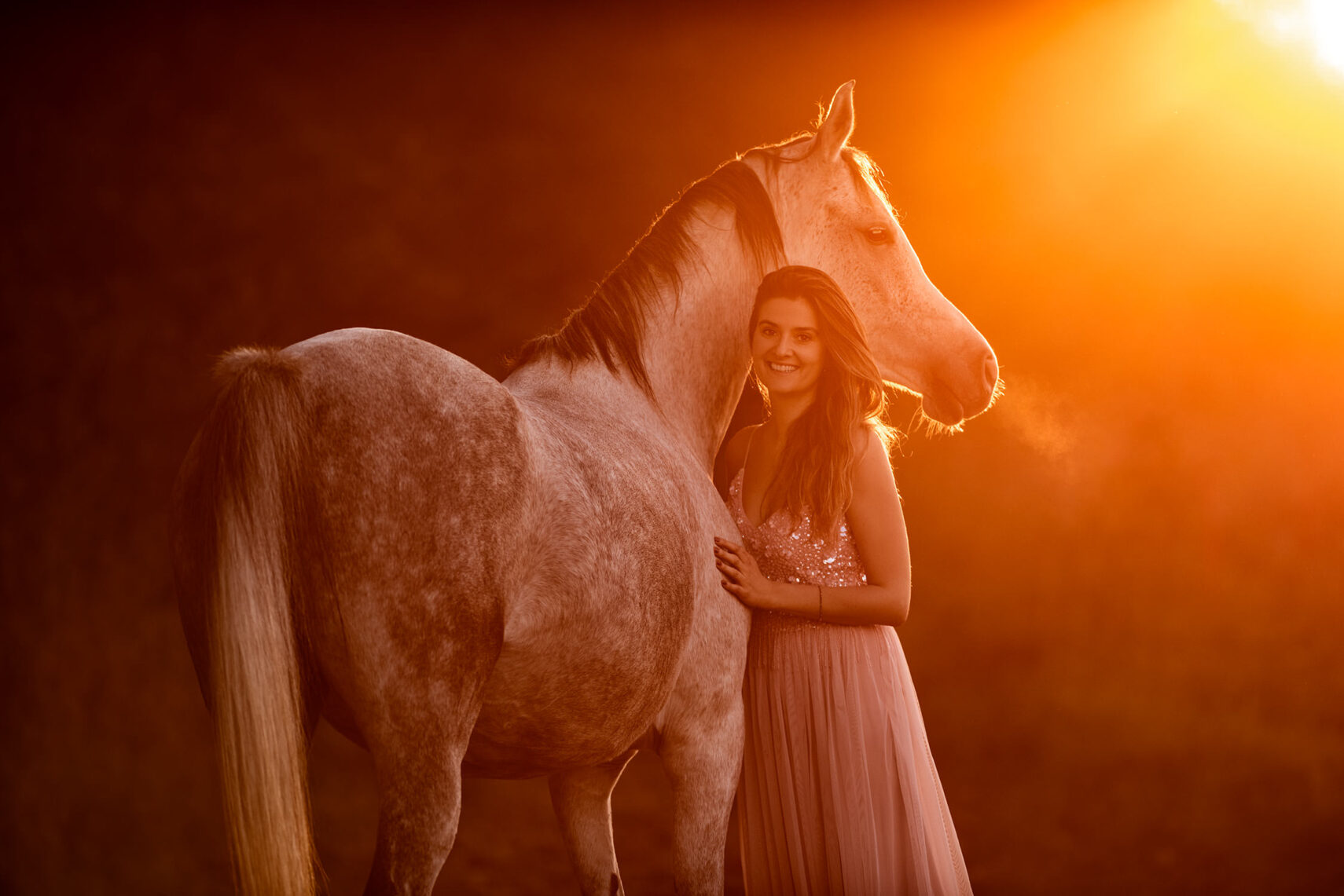 Eine Frau in einem glitzernden Kleid steht neben einem Pferd, beide blicken entspannt in die Kamera, umhüllt vom goldenen Licht der Sonne.