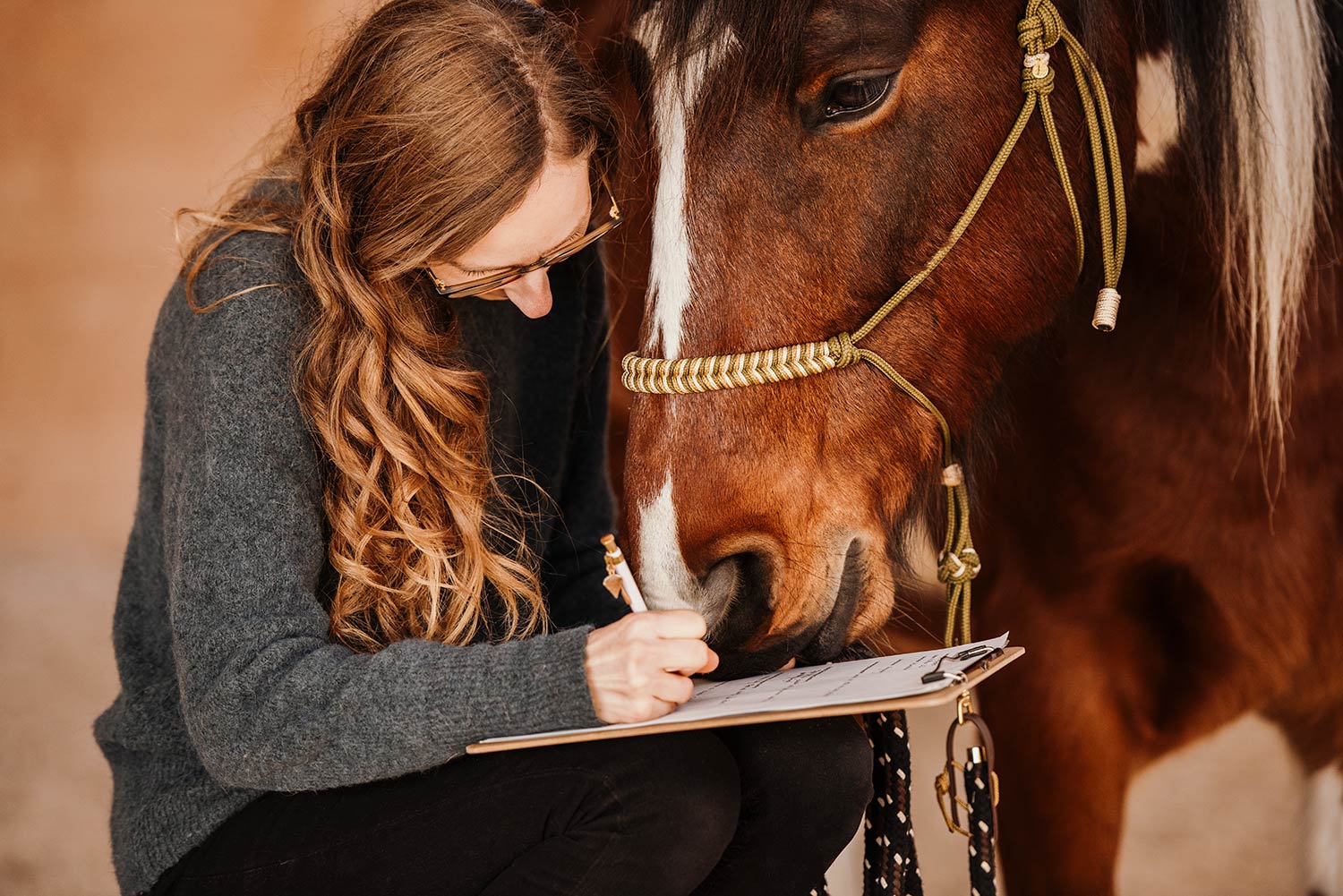 Eine Frau mit langen Haaren schreibt etwas auf einem Klemmbrett, während ein braunes Pferd danebensteht.