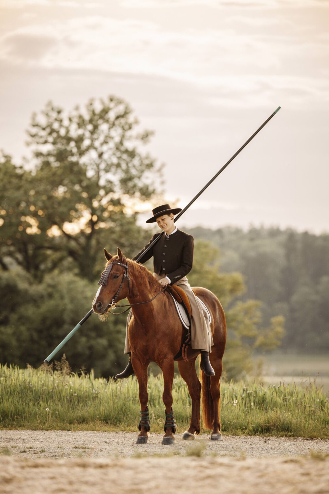 Eine Reiterin in traditioneller Kleidung reitet ihr braunes Pferd bei Sonnenuntergang und hält eine lange Stange.